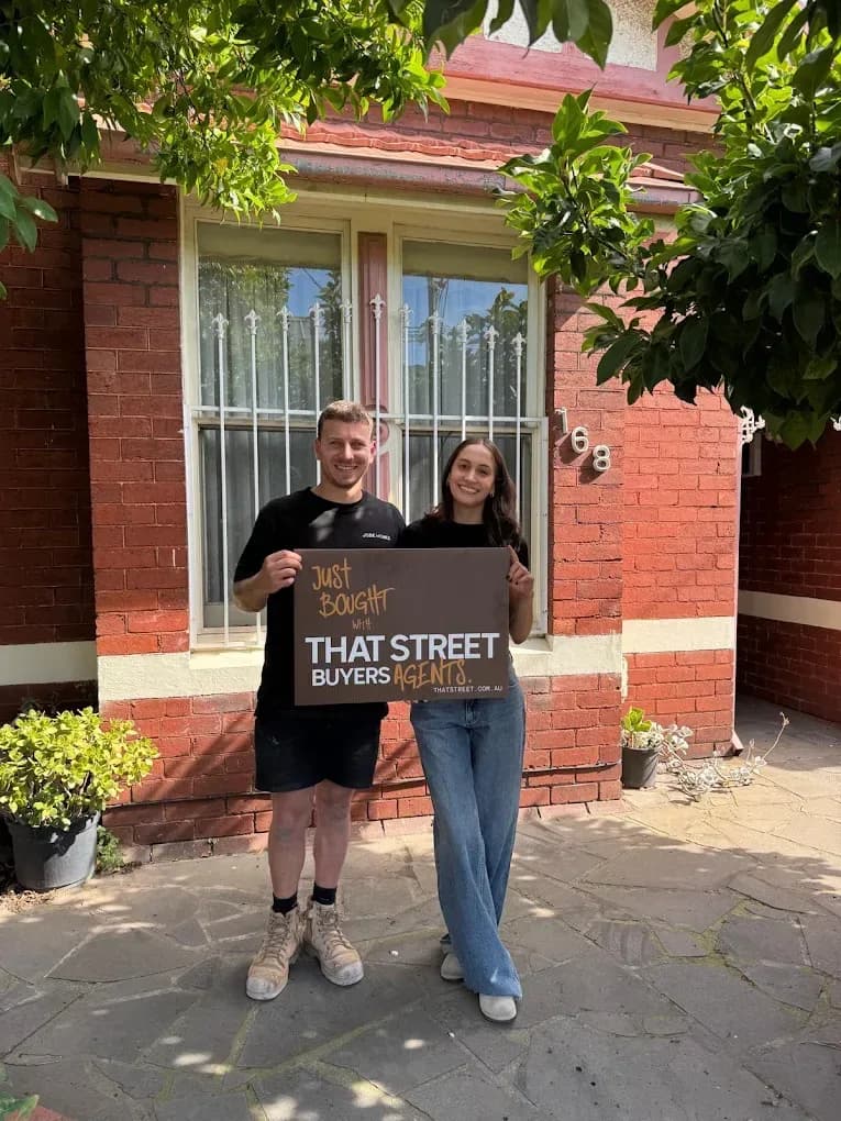 A couple outside their new red-brick home, holding a That Street buyers agent board between them