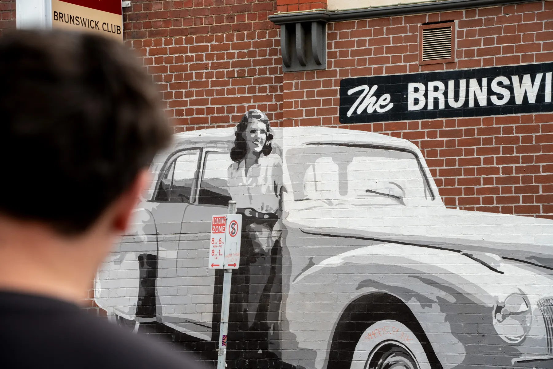 A painted mural of a woman beside a classic car on The Brunswick Club’s brick wall