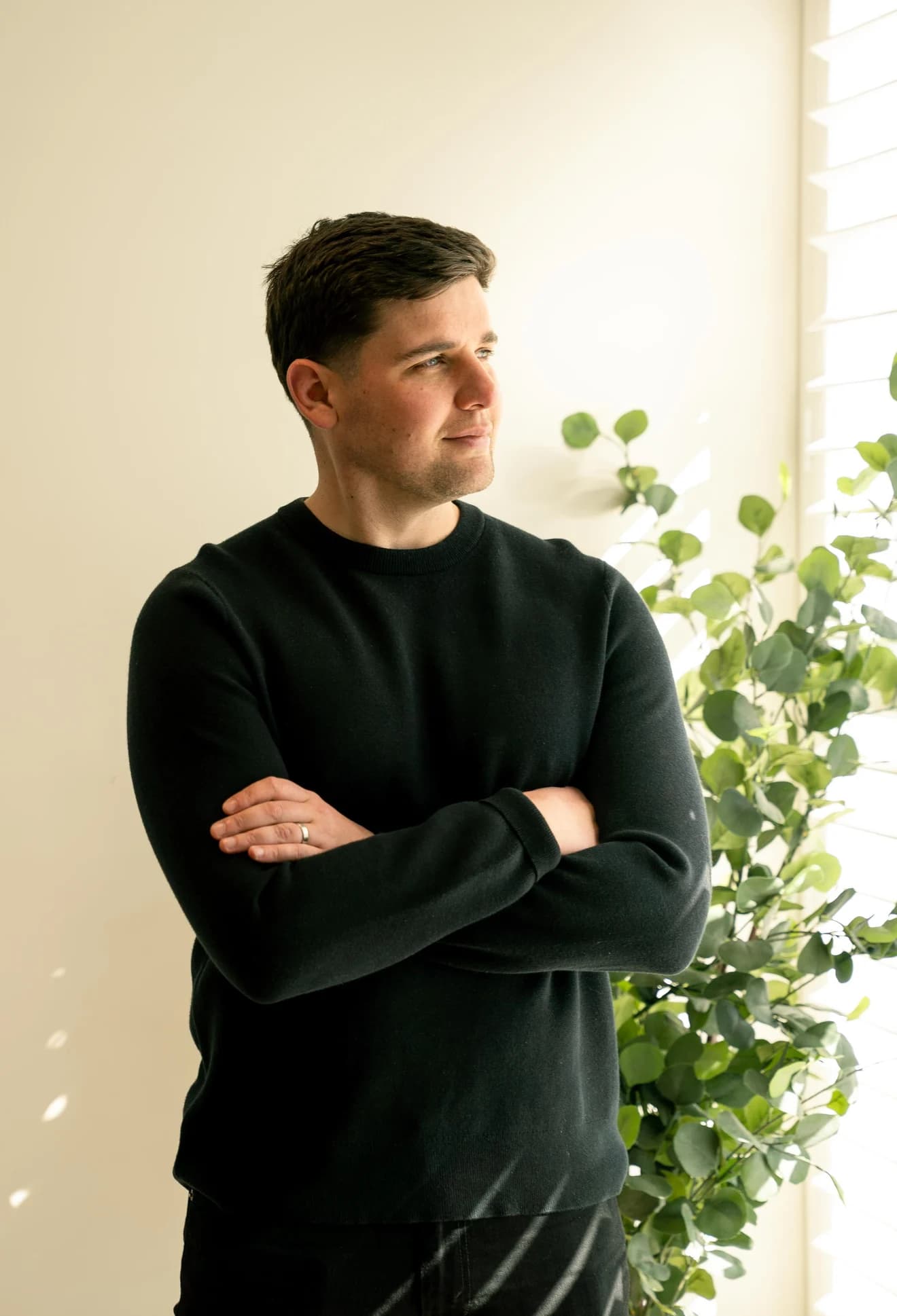 Jason Lekkas in a black jumper, arms folded by a shuttered window, a eucalypt beside him