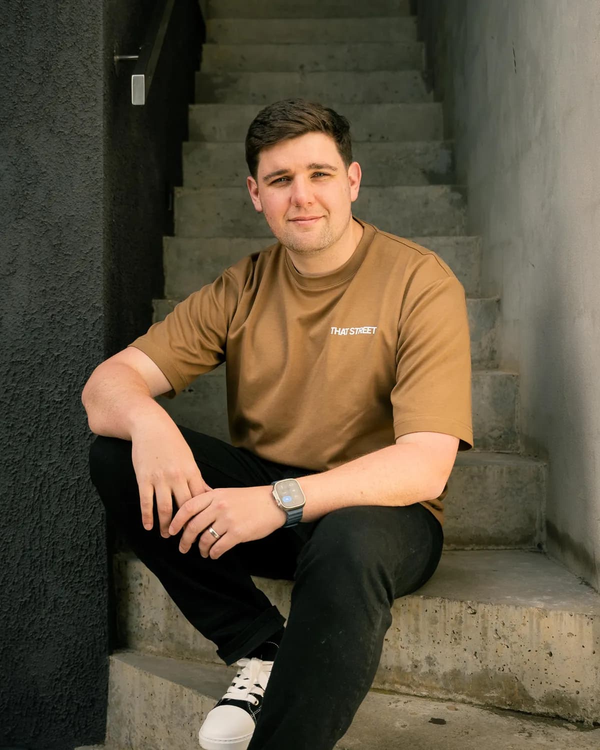 Jason Lekkas in a tan That Street tee, seated on concrete stairs between a dark wall and a sunlit one