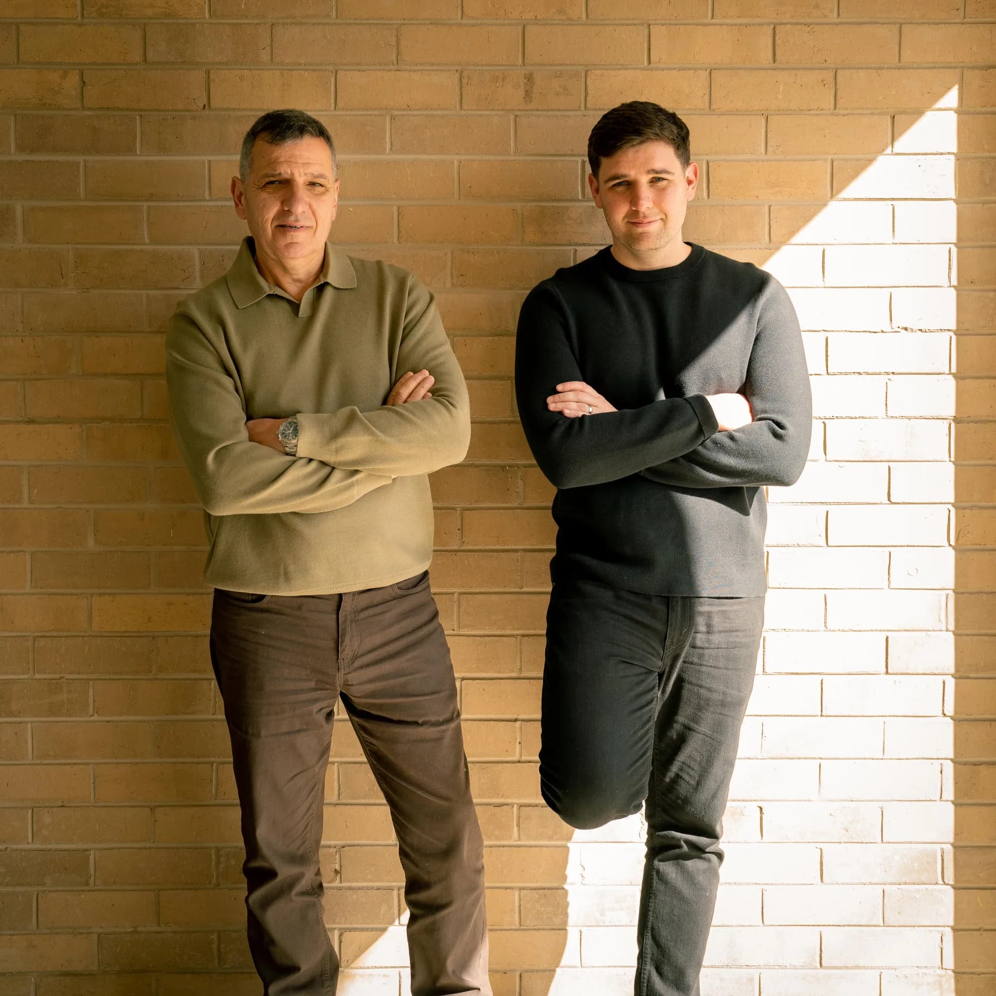 Jim and Jason Lekkas against a sunlit cream brick wall, arms folded — Jim on the left in an olive jumper, Jason on the right in black