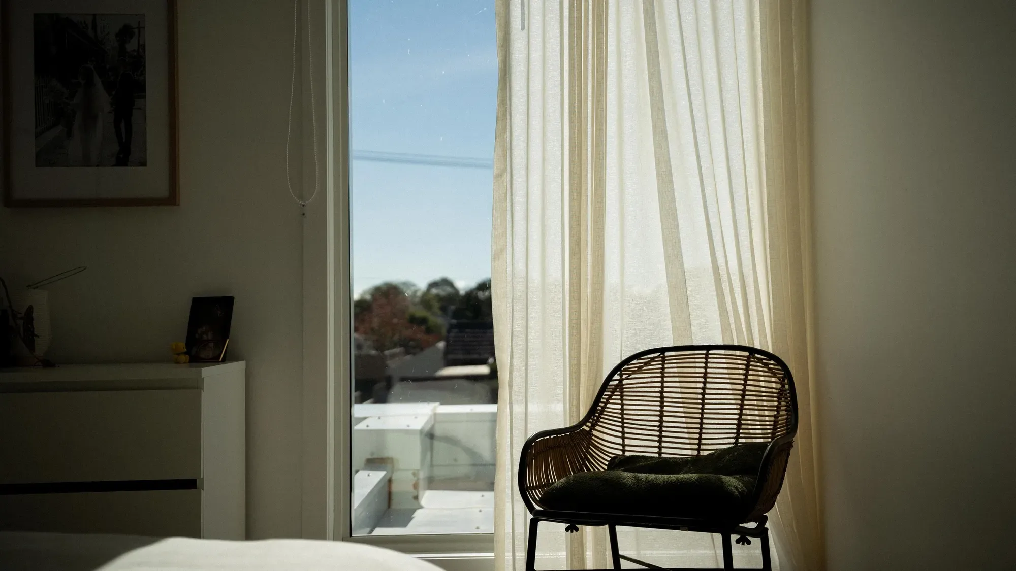 A bedroom with sheer curtains and a rattan chair by the window