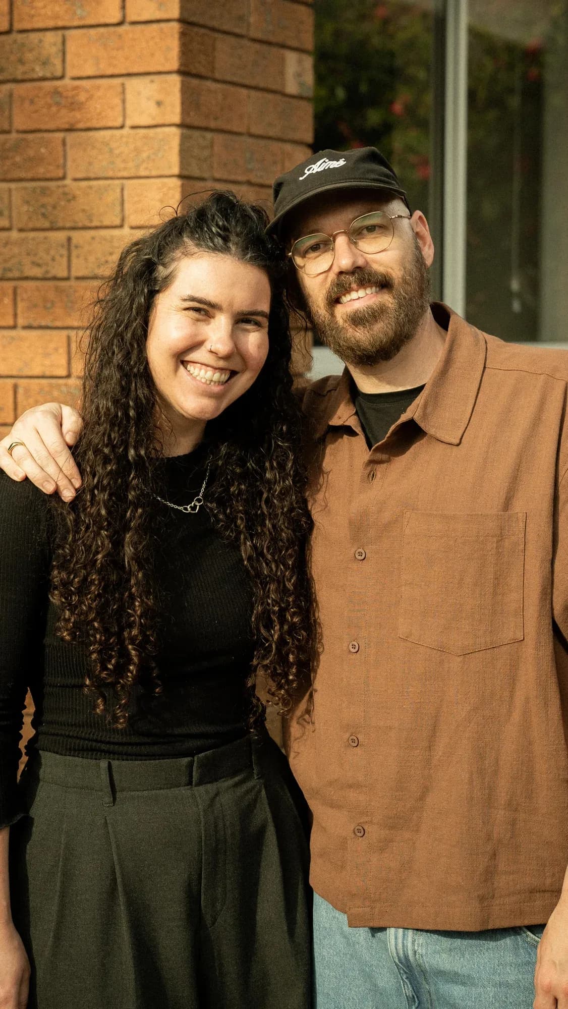 Dani and Shaughan against a red-brick wall, her in black, him in a rust shirt and cap