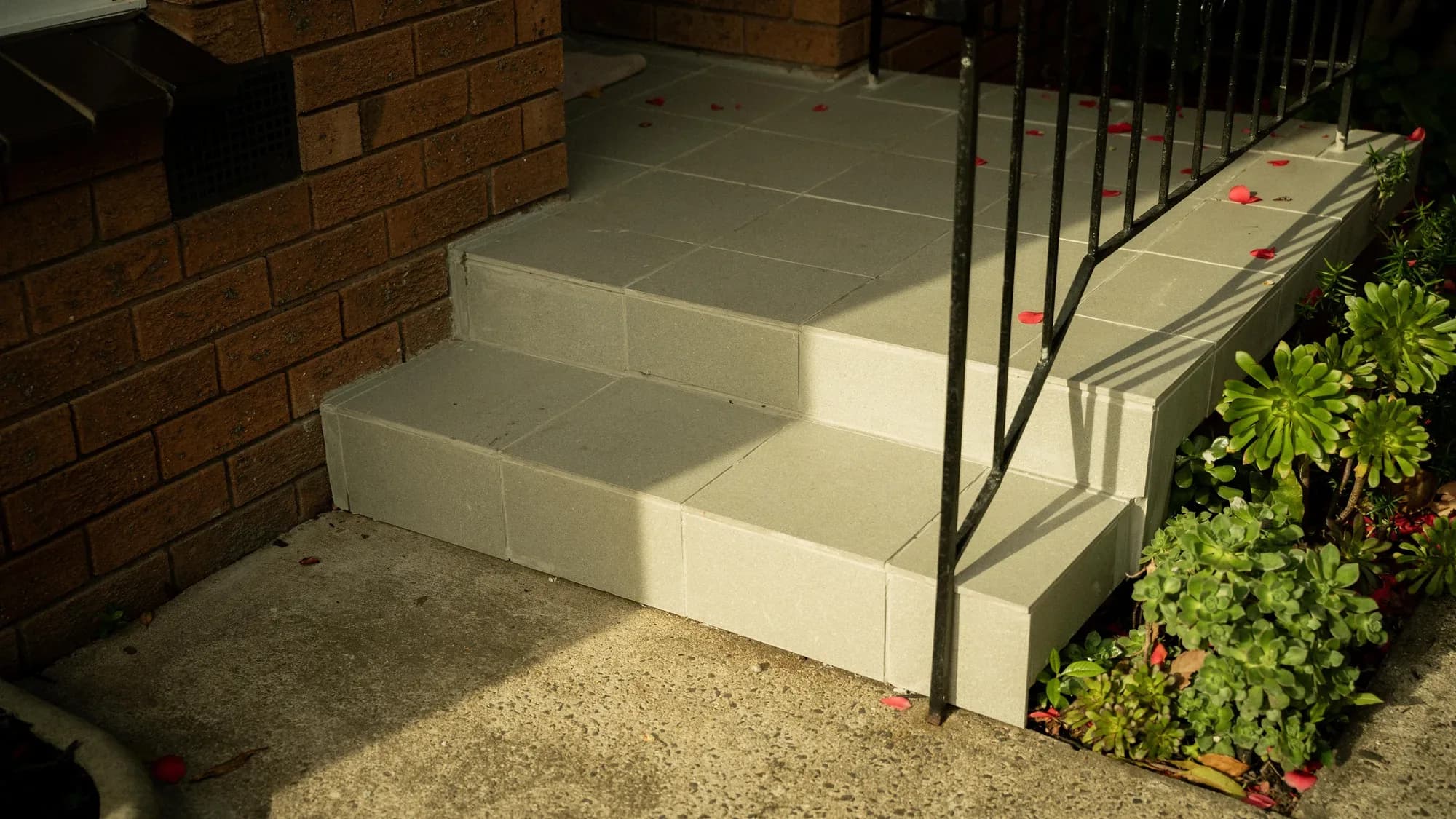 Three pale concrete front steps with a black rail, petals scattered on the treads