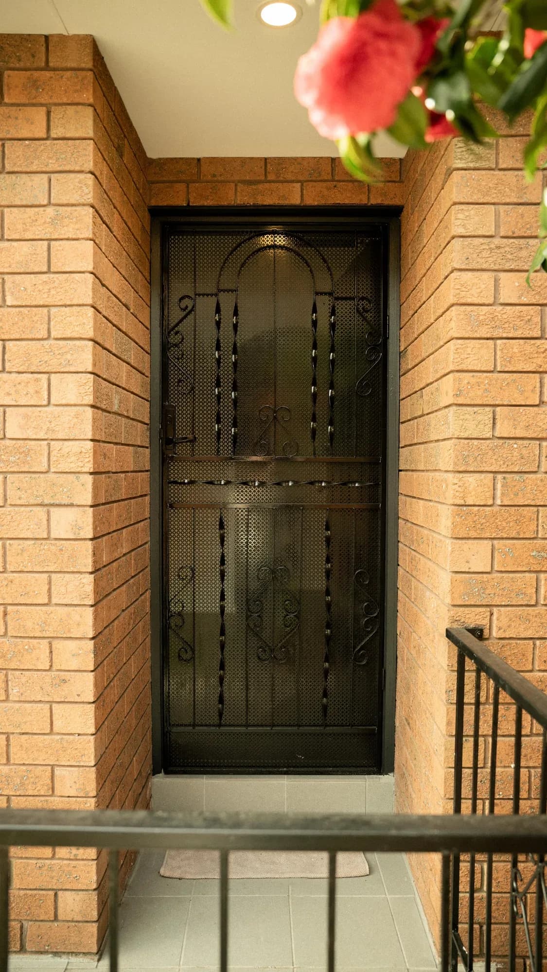 A black wrought-iron screen door set deep in a red-brick porch, camellias above